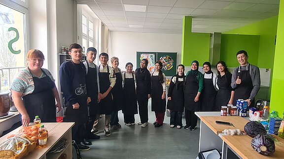 Usbekische Schülerinnen und Schüler stehen gemeinsam mit zwei Lehrkräften in der Lehrküche der Berufsschule Auerbach und bereiten regionale Gerichte zu.